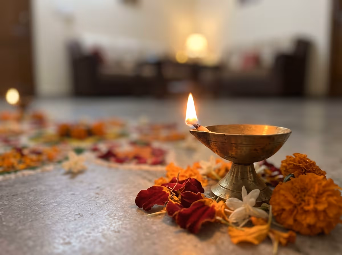 A lit brass oil lamp sits on a light-colored floor, surrounded by red, orange, and white flower petals.