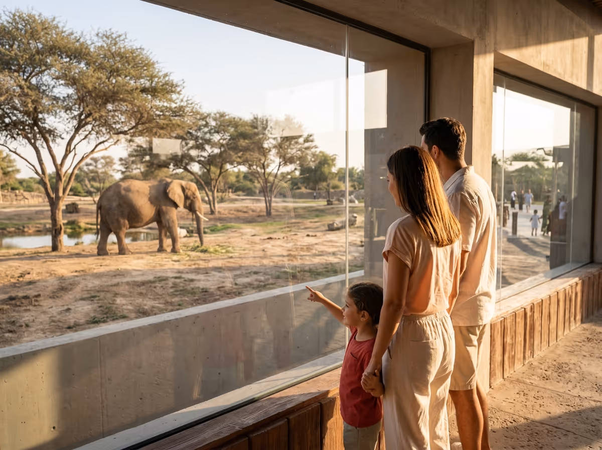 A family watches an elephant from behind a glass wall, with a child pointing at the animal in a sunny outdoor enclosure.