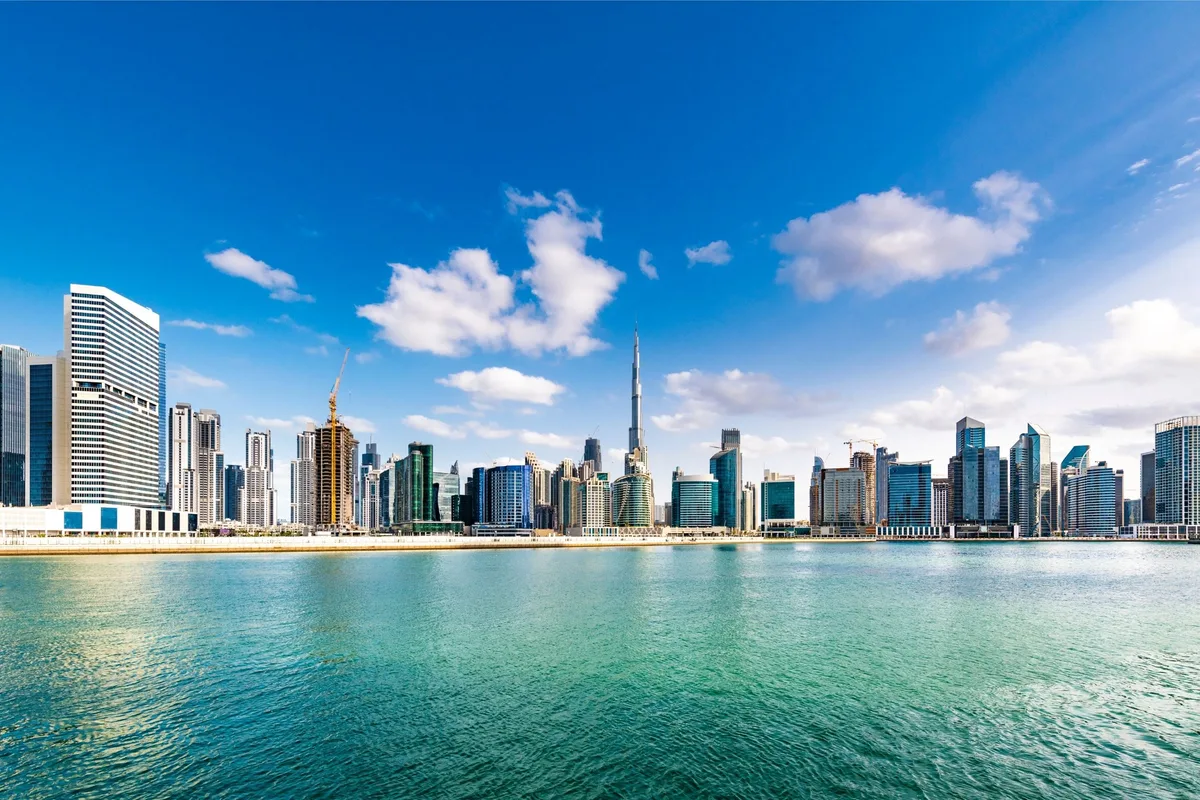 Skyline of Dubai with tall skyscrapers under a bright blue sky. Calm waterfront in the foreground.